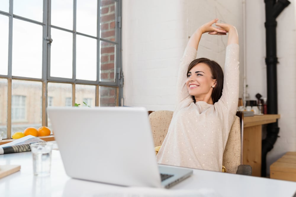 Happy relaxed young woman sitting in her kitchen with a laptop in front of her stretching her arms above her head and looking out of the window with a smile Happy relaxed young woman sitting in her kitchen with a laptop in front of her stretching her arms above her head and looking out of the window with a smile